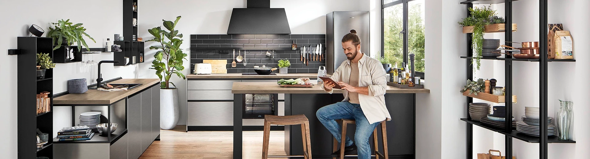 Junger Mann mit Tablet auf einem Barhocker in einer modernen Küche mit Edelstahlfronten und einer Nischenwandverkleidung in Fliesenoptik. Die Arbeitsplatte ist einem Holzton, Grünpflanzen ergänzen die Atmosphäre. Junger Mann mit Tablet auf einem Barhocker in einer modernen Küche mit Edelstahlfronten und einer Nischenwandverkleidung in Fliesenoptik. Die Arbeitsplatte ist einem Holzton, Grünpflanzen ergänzen die Atmosphäre.