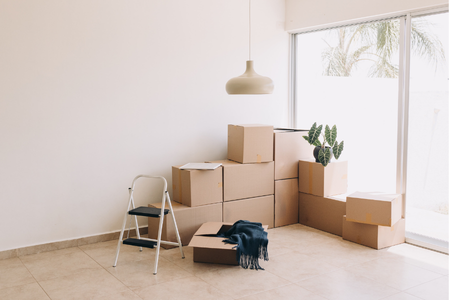 Leere Wohnungsecke mit gestapelten Umzugskartons, einer kleinen Leiter, einer Pflanze und einem Tuch auf dem Boden vor einer großen Fensterfront Leere Wohnungsecke mit gestapelten Umzugskartons, einer kleinen Leiter, einer Pflanze und einem Tuch auf dem Boden vor einer großen Fensterfront