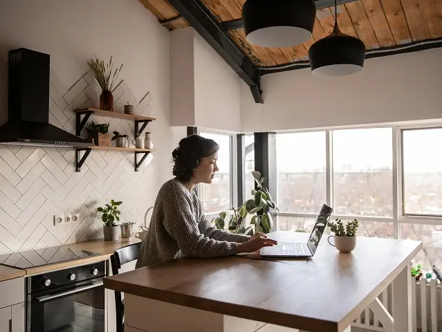Frau in einer gemütlichen Küche mit Holzdecke arbeitet an einem Holztisch an einem Laptop. Frau in einer gemütlichen Küche mit Holzdecke arbeitet an einem Holztisch an einem Laptop.