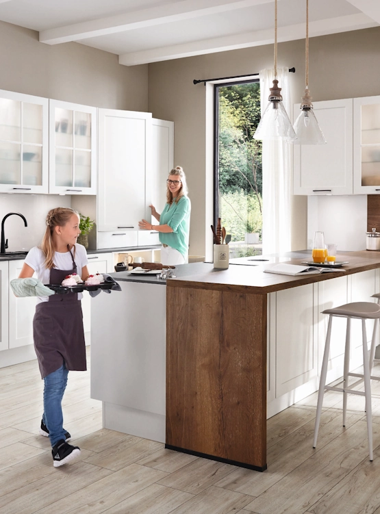 Eine Mutter und ihre Tochter backen gemeinsam in einer hellen, modernen Küche. Die Tochter trägt ein Backblech, während die Mutter im Hintergrund in einen Schrank schaut. Eine Mutter und ihre Tochter backen gemeinsam in einer hellen, modernen Küche. Die Tochter trägt ein Backblech, während die Mutter im Hintergrund in einen Schrank schaut.