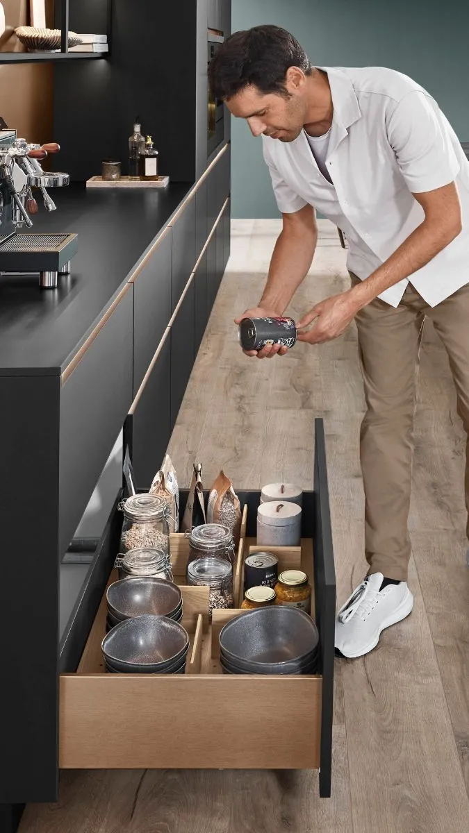 Ein Mann beugt sich über eine weit geöffnete, organisierte Schublade aus hellem Holz in einer modernen, schwarzen Küche. Ein Mann beugt sich über eine weit geöffnete, organisierte Schublade aus hellem Holz in einer modernen, schwarzen Küche.