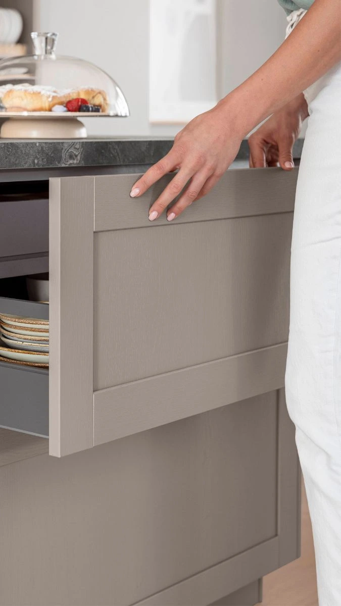 Eine Person zieht in einer Küche eine breite Schublade mit grauer, rahmenartigen Front heraus; darin sind gestapelte Teller zu sehen. Auf der dunklen Arbeitsplatte steht eine Glasglocke mit Gebäck. Eine Person zieht in einer Küche eine breite Schublade mit grauer, rahmenartigen Front heraus; darin sind gestapelte Teller zu sehen. Auf der dunklen Arbeitsplatte steht eine Glasglocke mit Gebäck.