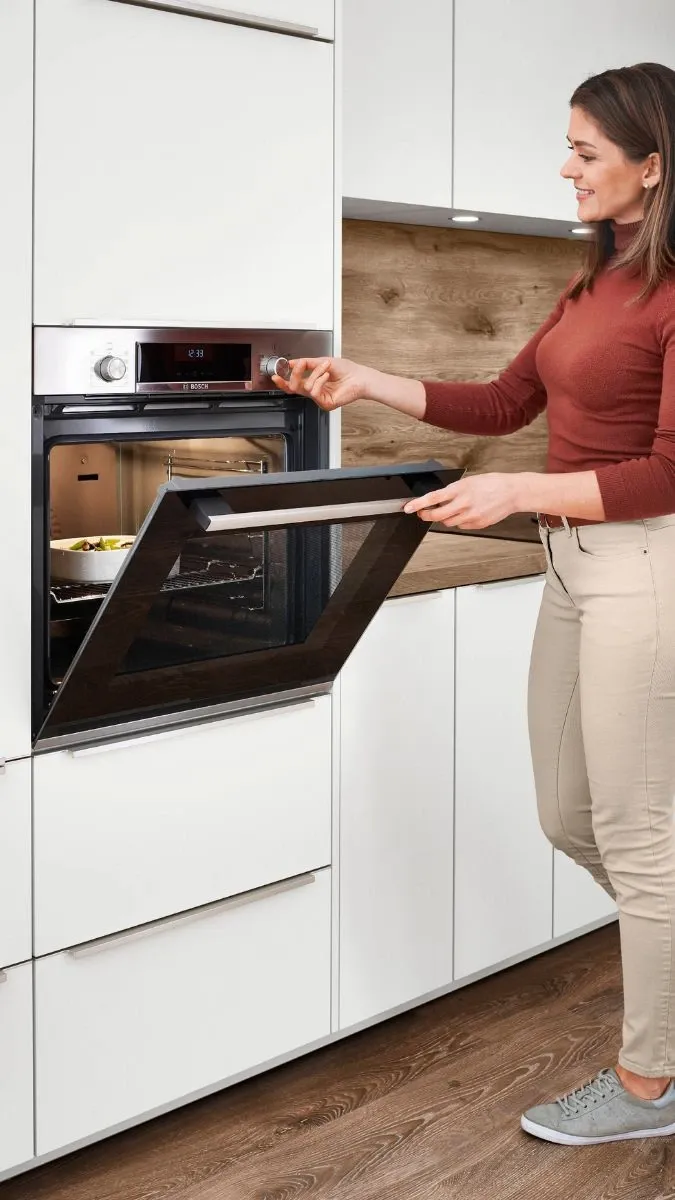 Eine Frau steht in einer modernen Küche, öffnet den Backofen, während sie mit der anderen Hand die Temperatur am Drehknopf einstellt Eine Frau steht in einer modernen Küche, öffnet den Backofen, während sie mit der anderen Hand die Temperatur am Drehknopf einstellt