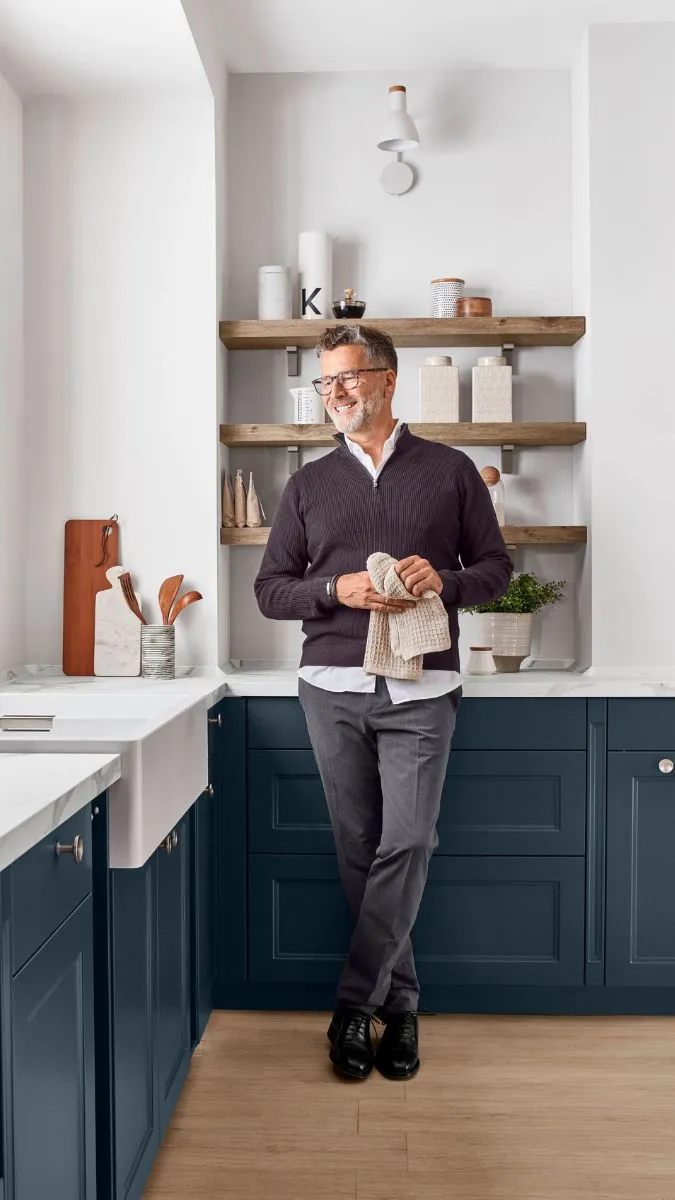 Lachender Mann im dunklen Pullover und Brille steht in einer modernen Küche mit dunkelblauen Schränken, weißer Spüle und offenen Holzregalen Lachender Mann im dunklen Pullover und Brille steht in einer modernen Küche mit dunkelblauen Schränken, weißer Spüle und offenen Holzregalen