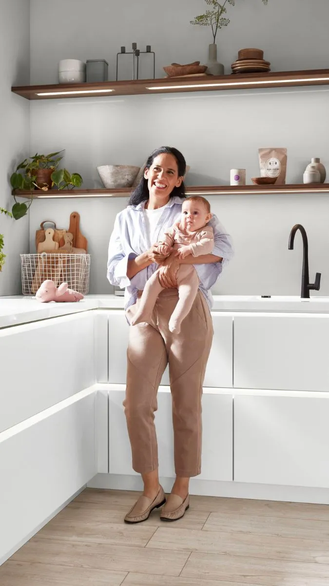 Helle, moderne Küche mit weißen Arbeitsplatten, Holzregalen und einer Frau, die ein Baby hält. Helle, moderne Küche mit weißen Arbeitsplatten, Holzregalen und einer Frau, die ein Baby hält.
