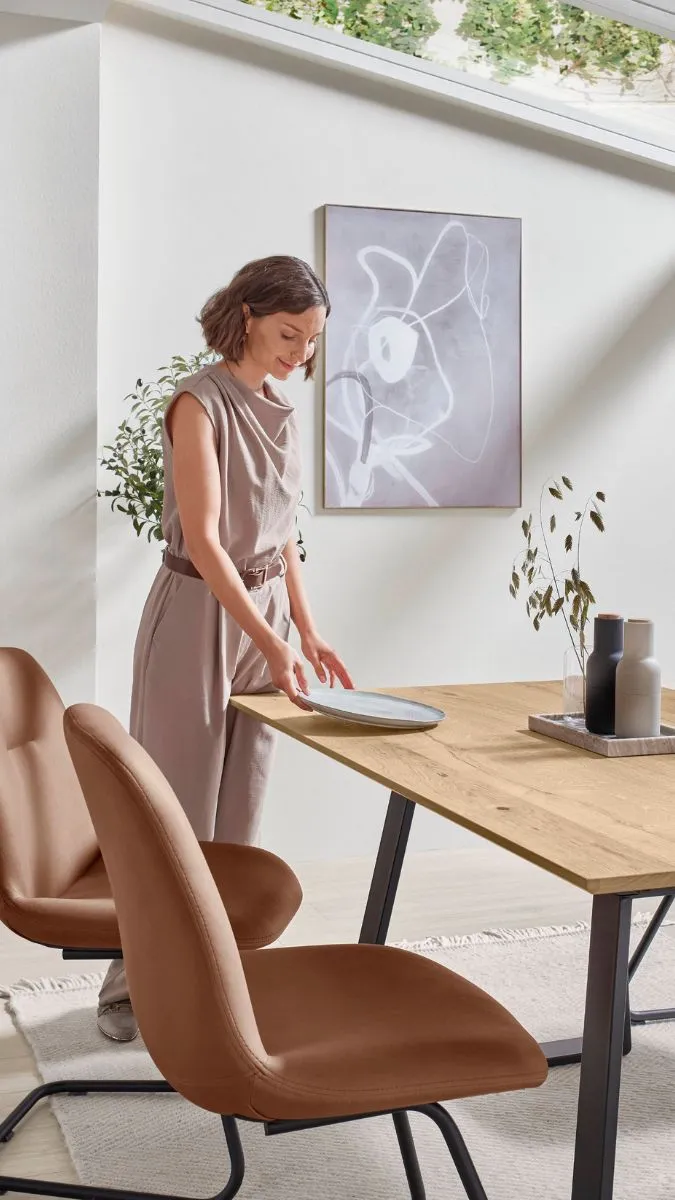 Frau in grauem Overall deckt Esstisch ein. Im Hintergrund hängt ein abstraktes Gemälde. Frau in grauem Overall deckt Esstisch ein. Im Hintergrund hängt ein abstraktes Gemälde.