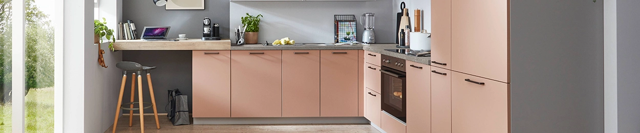 Moderne Küche mit roséfarbenen Fronten, dunklen Griffen, grauer Arbeitsplatte und integriertem Backofen; links ein kleiner Arbeitsplatz mit Holztheke und Hocker neben großen Fensterflächen. Moderne Küche mit roséfarbenen Fronten, dunklen Griffen, grauer Arbeitsplatte und integriertem Backofen; links ein kleiner Arbeitsplatz mit Holztheke und Hocker neben großen Fensterflächen.