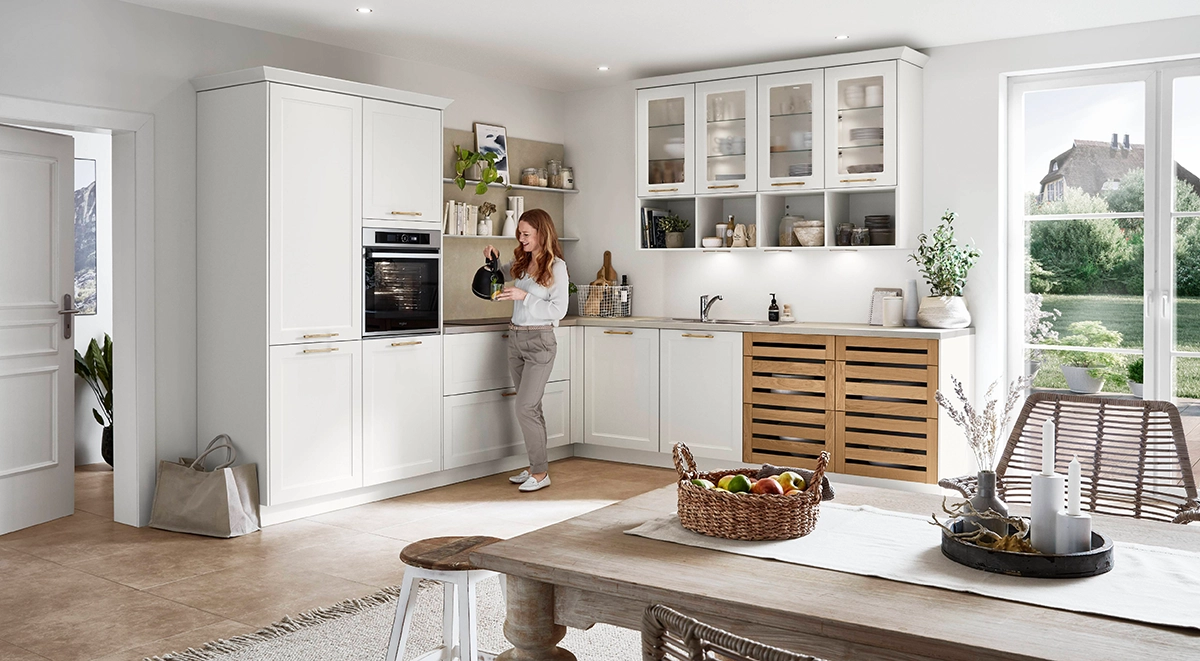 Helle Küche im Landhausstil mit weißen Fronten, hohen Schränken und Glastüren an den Oberschränken; rechts Schubladen im Holz-Lamellendesign, große Fenster mit Gartenblick und ein gemütlicher Essbereich im Vordergrund. Helle Küche im Landhausstil mit weißen Fronten, hohen Schränken und Glastüren an den Oberschränken; rechts Schubladen im Holz-Lamellendesign, große Fenster mit Gartenblick und ein gemütlicher Essbereich im Vordergrund.