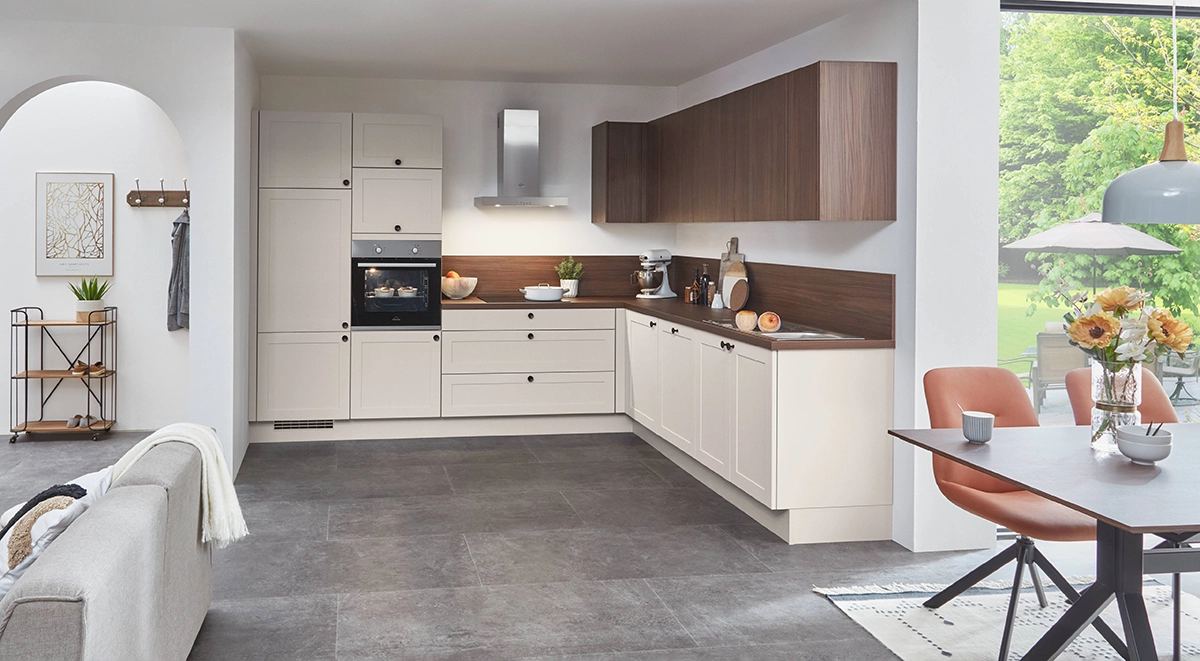 Helle L-Küche mit cremefarbenen Fronten, dunkler Holzarbeitsplatte und passenden Oberschränken; integrierter Backofen und angrenzender Essbereich mit großen Fensterflächen. Helle L-Küche mit cremefarbenen Fronten, dunkler Holzarbeitsplatte und passenden Oberschränken; integrierter Backofen und angrenzender Essbereich mit großen Fensterflächen.