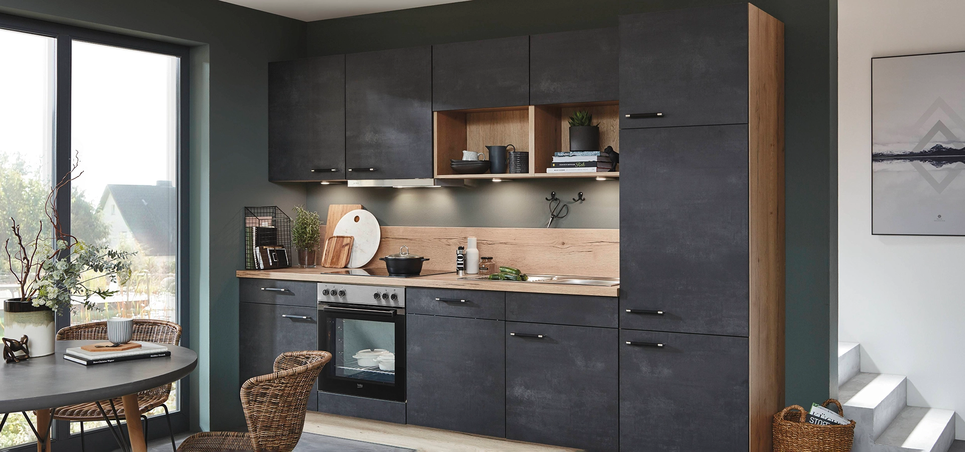 Wir sehen eine einzeilige Küche mit Front in schwarzbeton und der Arbeitsplatte in einem Holzdekor. Davor steht ein Esstisch mit Korbstühlen. Wir sehen eine einzeilige Küche mit Front in schwarzbeton und der Arbeitsplatte in einem Holzdekor. Davor steht ein Esstisch mit Korbstühlen.