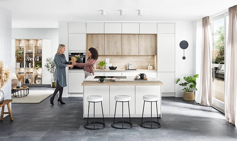 Zwei junge Frauen treffen sich in einer neuen offenen Küche mit weißen glatten Fronten. Die Küche ist grifflos und besteht aus einer Zeile mit Hochschränken sowie einer Insel mit Barhockern. Zwei junge Frauen treffen sich in einer neuen offenen Küche mit weißen glatten Fronten. Die Küche ist grifflos und besteht aus einer Zeile mit Hochschränken sowie einer Insel mit Barhockern.