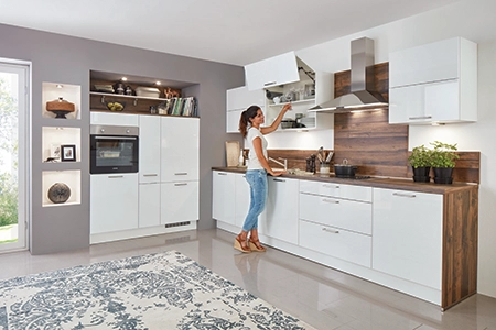 Eine junge Frau in einer weißen Küche mit Arbeitsplatte in Holzton putzt beim Frühjahrsputz von oben nach unten. Gerade öffnet sie einen Faltsliftwandschrank. Eine junge Frau in einer weißen Küche mit Arbeitsplatte in Holzton putzt beim Frühjahrsputz von oben nach unten. Gerade öffnet sie einen Faltsliftwandschrank.