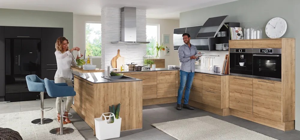 Helle Wohnküche in Holzoptik mit Kochinsel und Sitzplätzen, zwei Personen bereiten gemeinsam Essen zu, integrierte Einbaugeräte, Dunstabzug über dem Kochfeld und moderne, wohnliche Gestaltung. Helle Wohnküche in Holzoptik mit Kochinsel und Sitzplätzen, zwei Personen bereiten gemeinsam Essen zu, integrierte Einbaugeräte, Dunstabzug über dem Kochfeld und moderne, wohnliche Gestaltung.