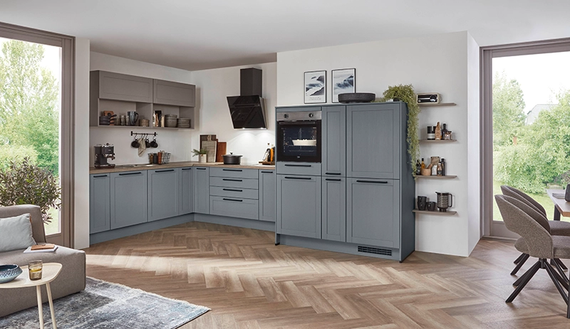 Rauchblaue Küche mit Holzmaserung. Die Küche hat eine L-Form und drei Highboards. Bodenbelag mit Fischgrätmuster, rechts und links zwei bodentiefe Fenster. Rauchblaue Küche mit Holzmaserung. Die Küche hat eine L-Form und drei Highboards. Bodenbelag mit Fischgrätmuster, rechts und links zwei bodentiefe Fenster.