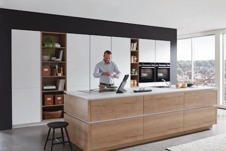 Moderne Wohnküche mit großer Kochinsel aus Holz und weißer Arbeitsplatte, Mann bereitet Essen zu, im Hintergrund Hochschränke mit Einbaugeräten und große Fensterfront mit Tageslicht. Moderne Wohnküche mit großer Kochinsel aus Holz und weißer Arbeitsplatte, Mann bereitet Essen zu, im Hintergrund Hochschränke mit Einbaugeräten und große Fensterfront mit Tageslicht.