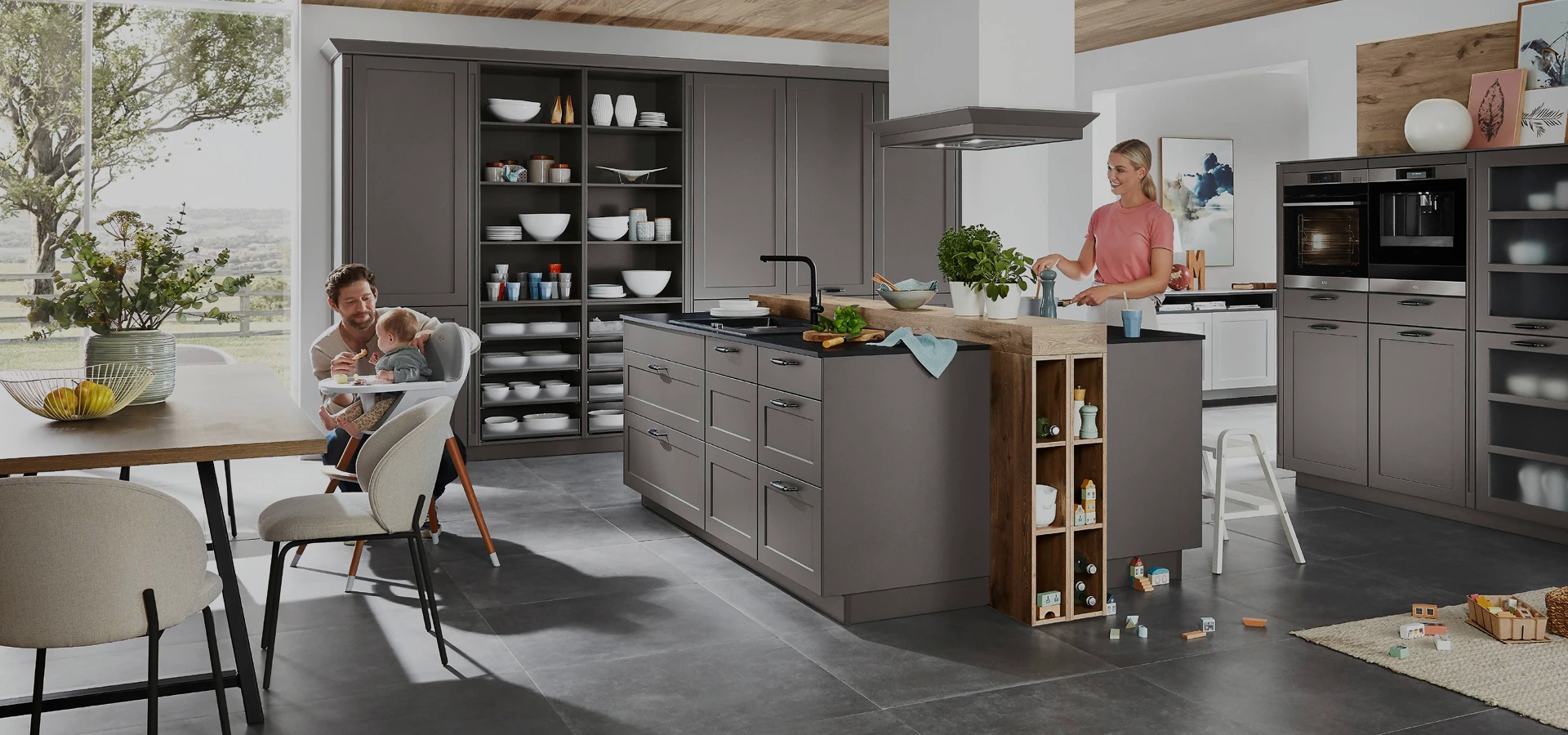 In einer hellen, modernen Küche mit grauen Schränken, einer großen Kochinsel, offenen Regalen und Holzakzenten bereitet eine Frau an der Kochinsel Essen zu, während ein Mann mit einem Kind am Esstisch sitzt. In einer hellen, modernen Küche mit grauen Schränken, einer großen Kochinsel, offenen Regalen und Holzakzenten bereitet eine Frau an der Kochinsel Essen zu, während ein Mann mit einem Kind am Esstisch sitzt.