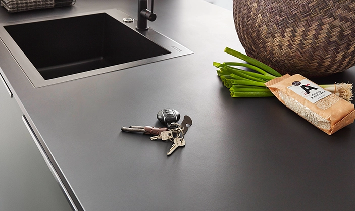 Blick auf eine Arbeitsplatte in betongrau. Man sieht eine nahtlos eingebaute Spüle. Schlüssel sowie Gemüse und Einkaufskorb liegen auf der Arbeitsplatte. Blick auf eine Arbeitsplatte in betongrau. Man sieht eine nahtlos eingebaute Spüle. Schlüssel sowie Gemüse und Einkaufskorb liegen auf der Arbeitsplatte.