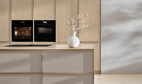 Wir sehen eine Japandi-Küche mit Hochschrankzeile und Kochinsel. Die Hochschränke haben einen Holzton, die Insel hat eine hellbraune Front. Wir sehen eine Japandi-Küche mit Hochschrankzeile und Kochinsel. Die Hochschränke haben einen Holzton, die Insel hat eine hellbraune Front.