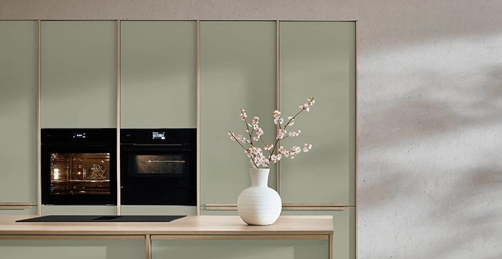 Moderne Küche mit matten, grünen Fronten, zwei schwarzen Einbaugeräten und einer hellen Arbeitsfläche, auf der eine weiße Vase mit Blüten steht. Moderne Küche mit matten, grünen Fronten, zwei schwarzen Einbaugeräten und einer hellen Arbeitsfläche, auf der eine weiße Vase mit Blüten steht.