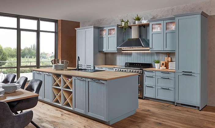 Eine Küche, die Trends mit Landhhausstil vereint: Steinblaue Rahmenfronten mit Holzstruktur. Eine Zeile sowie eine Insel. Im Vordergrund links ein Tisch. Eine Küche, die Trends mit Landhhausstil vereint: Steinblaue Rahmenfronten mit Holzstruktur. Eine Zeile sowie eine Insel. Im Vordergrund links ein Tisch.