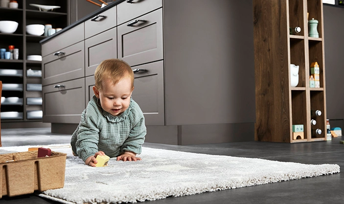 Ein Baby krabbelt auf einem Teppich in einer modernen Landhausküche mit grauen matten Rahmenfronten. Ein Baby krabbelt auf einem Teppich in einer modernen Landhausküche mit grauen matten Rahmenfronten.