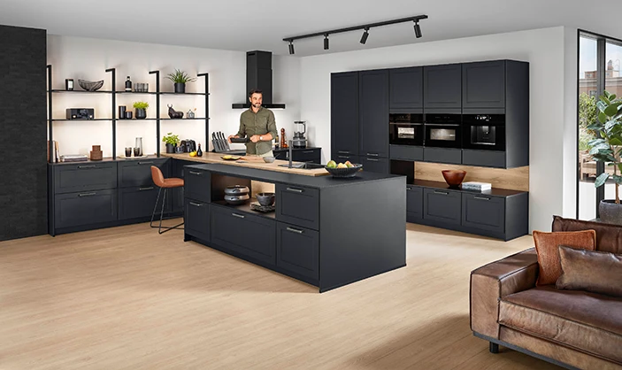 Ein Mann kocht in einer großen modernen Landhausküche mit schwarzen matten Fronten. Links Unterschränke mit Regalen, rechts eine Hochschrankzeile und zentral eine Kochinsel. Ein Mann kocht in einer großen modernen Landhausküche mit schwarzen matten Fronten. Links Unterschränke mit Regalen, rechts eine Hochschrankzeile und zentral eine Kochinsel.