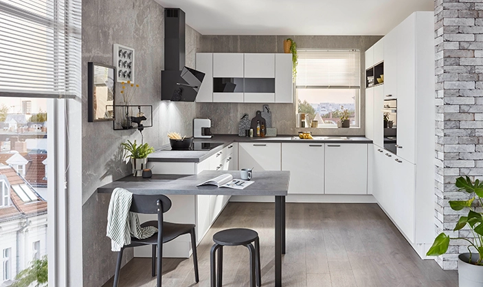 Weiße Küche in U-Form mit einer Hochschrankzeile auf der rechten Seite und dunkler Arbeitsplatte. Vor der Küche steht ein Esstisch mit Stuhl und Hocker. Weiße Küche in U-Form mit einer Hochschrankzeile auf der rechten Seite und dunkler Arbeitsplatte. Vor der Küche steht ein Esstisch mit Stuhl und Hocker.