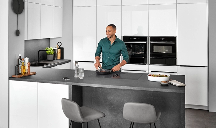 Junger Mann in grünem Hemd steht in einer U-Küche mit weißen Hochglanzfronten. Die Arbeitsplatte ist betongrau. Junger Mann in grünem Hemd steht in einer U-Küche mit weißen Hochglanzfronten. Die Arbeitsplatte ist betongrau.