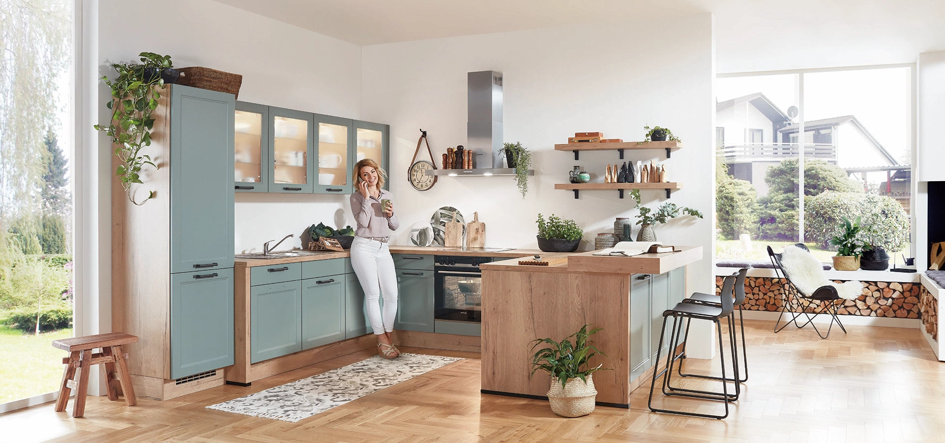 Moderne U-Küche mit mintfarbenen Fronten, Holzelementen, Kochinsel und großen Fenstern mit Blick ins Grüne; eine Person steht in der Küche und hält ein Getränk. Moderne U-Küche mit mintfarbenen Fronten, Holzelementen, Kochinsel und großen Fenstern mit Blick ins Grüne; eine Person steht in der Küche und hält ein Getränk.