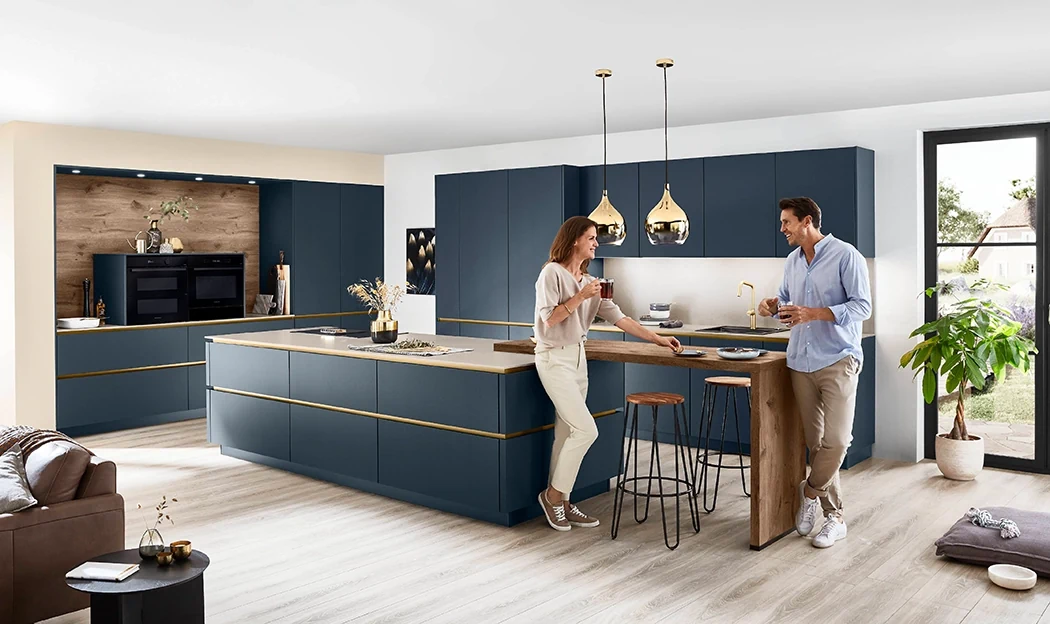 Ein Paar steht in einer individuell geplanten Küche mit glatten dunkelblauen Frontne und goldener Griffleiste. Es gibt zwei Zeilen sowie eine Kochinsel. Sie stehen an einer Theke mit Barhockern. Ein Paar steht in einer individuell geplanten Küche mit glatten dunkelblauen Frontne und goldener Griffleiste. Es gibt zwei Zeilen sowie eine Kochinsel. Sie stehen an einer Theke mit Barhockern.