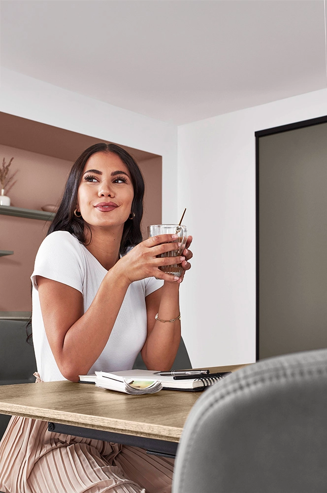 Junge Dame trinkt Kaffee am Küchentisch. Hinter ihr sehen wir ein graues Küchenregal mit Dekoobjekten.