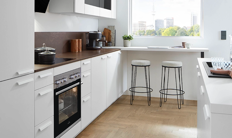 Küchen-Set, das man online bestellen kann. Weiße matte Fronten, weiße Griffe und Arbeitsplatte in einem Holzton. Rechts von der Küchenzeile ein Fenster mit kleiner Theke und zwei Barhockern. Küchen-Set, das man online bestellen kann. Weiße matte Fronten, weiße Griffe und Arbeitsplatte in einem Holzton. Rechts von der Küchenzeile ein Fenster mit kleiner Theke und zwei Barhockern.