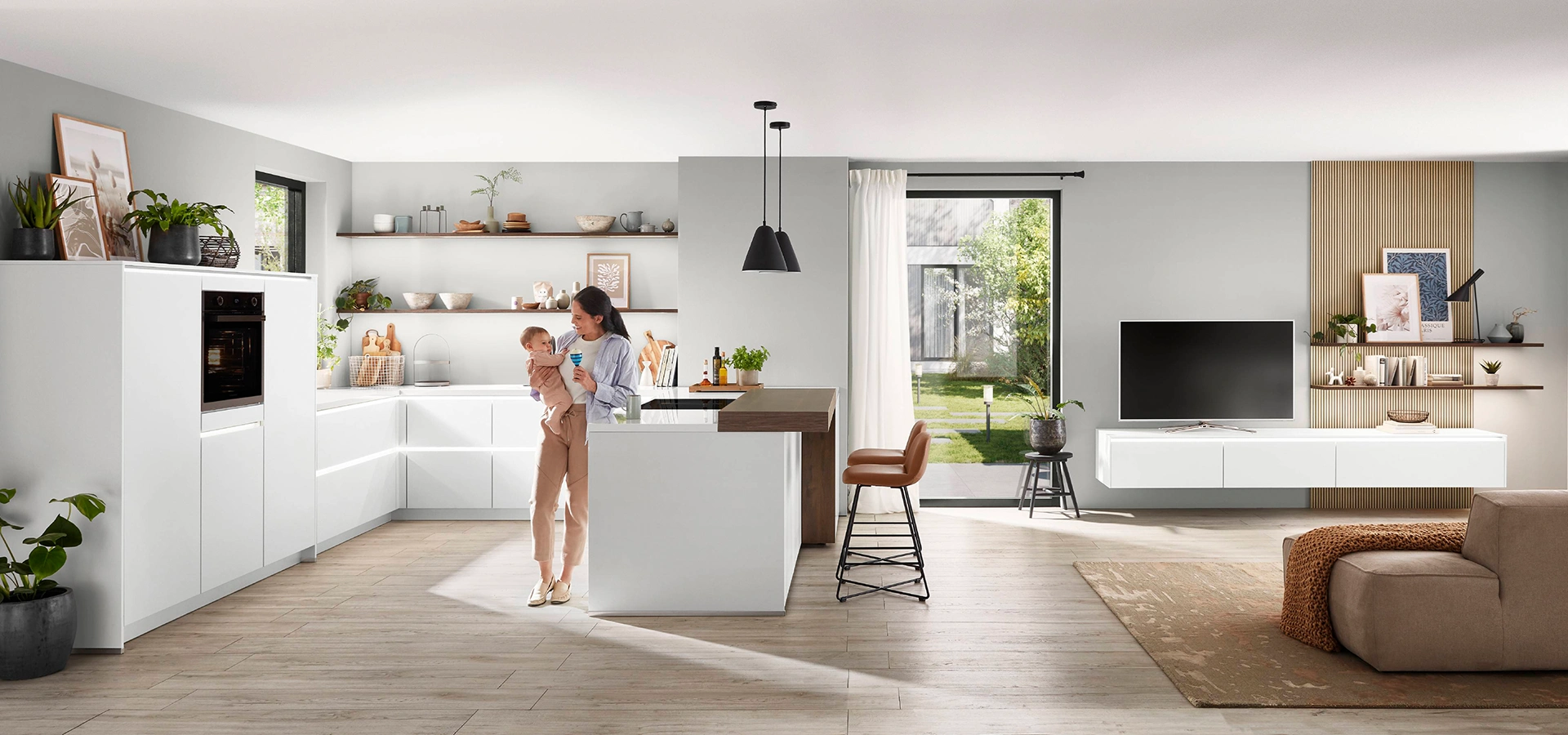 Eine junge Frau mit Baby steht in einer weißen offenen grifflosen Küche. Rechts sehen wir das Wohnzimmer. Eine junge Frau mit Baby steht in einer weißen offenen grifflosen Küche. Rechts sehen wir das Wohnzimmer.