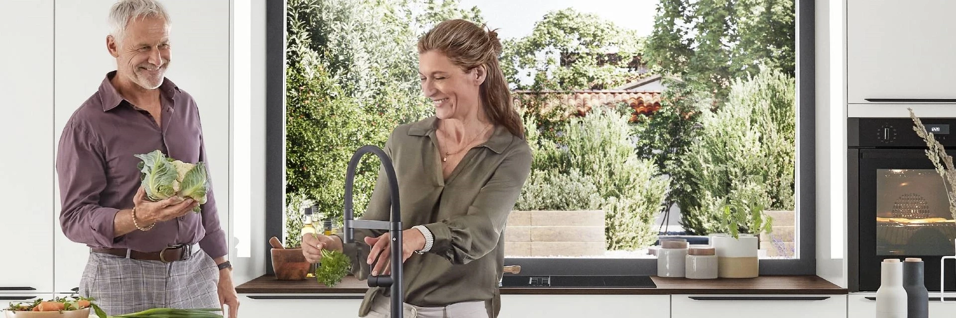 Moderne helle Küche mit Kücheninsel, großen Stauraum-Elementen und Panoramafenster – glückliches Paar bereitet gemeinsam frisches Gemüse zu