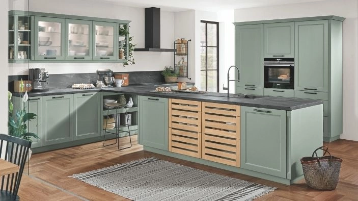 Landhausküche in sanftem Salbeigrün mit Rahmenfronten, großer Insel mit dunkler Arbeitsplatte und mittleren Lamellentüren in Holz, Glasoberschränke, offene Regale, integrierter Backofen, Kochfeld mit Wandhaube, Spülbecken mit hoher Armatur. Landhausküche in sanftem Salbeigrün mit Rahmenfronten, großer Insel mit dunkler Arbeitsplatte und mittleren Lamellentüren in Holz, Glasoberschränke, offene Regale, integrierter Backofen, Kochfeld mit Wandhaube, Spülbecken mit hoher Armatur.