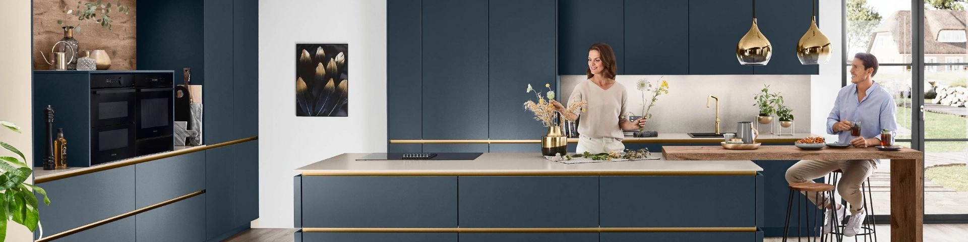 Moderne Küche in tiefblau mit goldenen Akzenten, großer Kücheninsel mit hellem Arbeitsplattenbelag, integrierten schwarzen Einbaugeräten, goldfarbenen Pendelleuchten, Holz-Stehtisch mit Barhockern. Moderne Küche in tiefblau mit goldenen Akzenten, großer Kücheninsel mit hellem Arbeitsplattenbelag, integrierten schwarzen Einbaugeräten, goldfarbenen Pendelleuchten, Holz-Stehtisch mit Barhockern.