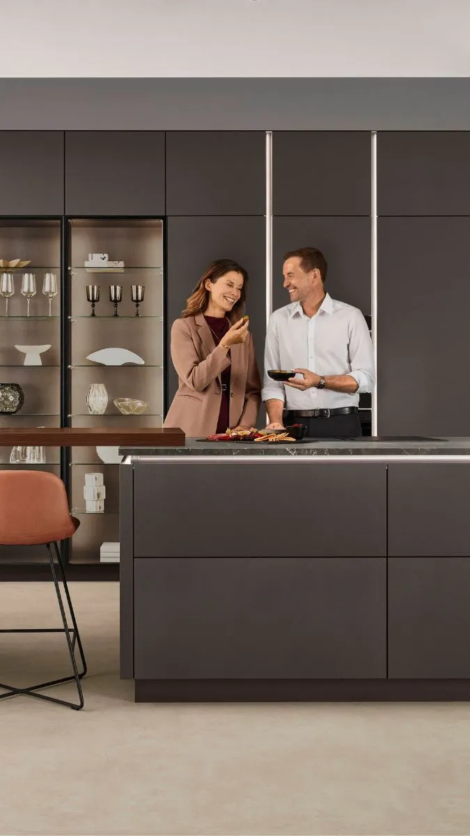 Moderne Küche in dunklem Braun mit Kücheninsel; zwei Personen stehen entspannt davor und unterhalten sich, im Hintergrund ein Glasvitrinenschrank mit Geschirr. Moderne Küche in dunklem Braun mit Kücheninsel; zwei Personen stehen entspannt davor und unterhalten sich, im Hintergrund ein Glasvitrinenschrank mit Geschirr.