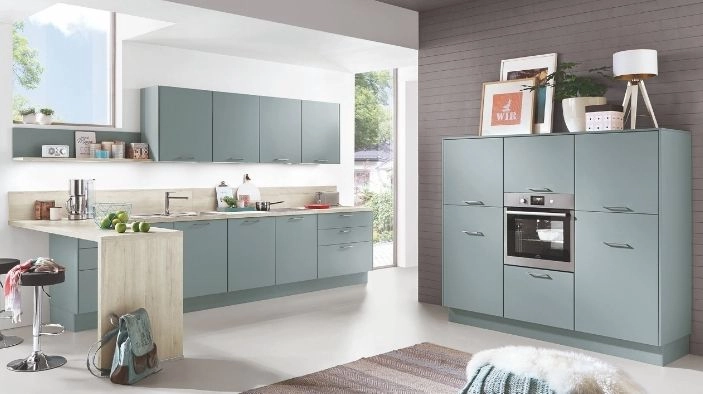 Moderne Einbauküche mit matten Fronten in Blaugrün, heller Arbeitsplatte, Hochschrank mit integriertem Backofen und kleiner Kücheninsel am Fenster. Moderne Einbauküche mit matten Fronten in Blaugrün, heller Arbeitsplatte, Hochschrank mit integriertem Backofen und kleiner Kücheninsel am Fenster.