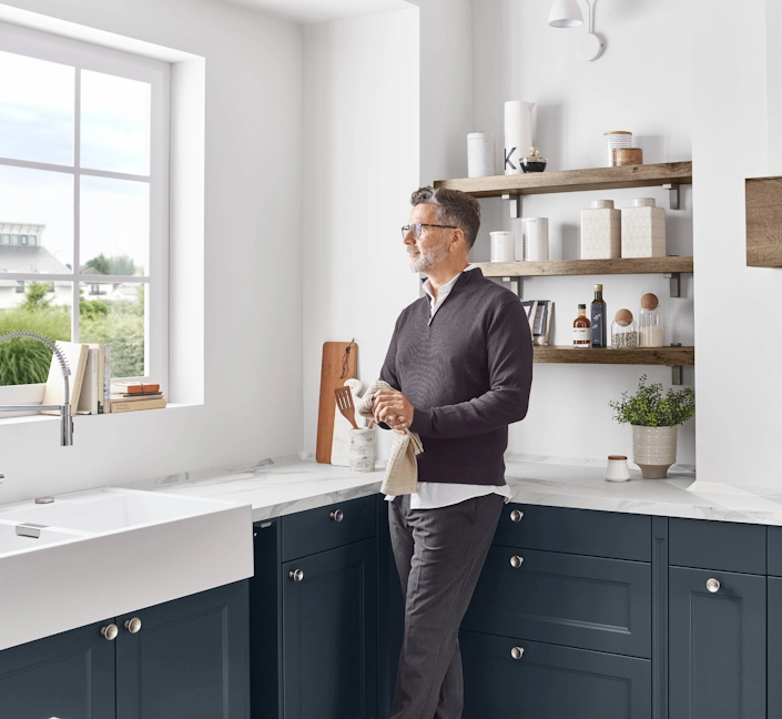 Ein Mann steht in einer modernen Küche neben der Spüle und trocknet Geschirr ab. Er schaut ruhig aus dem Fenster. Im Hintergrund befinden sich offene Holzregale mit Vorratsbehältern und Küchenutensilien. Ein Mann steht in einer modernen Küche neben der Spüle und trocknet Geschirr ab. Er schaut ruhig aus dem Fenster. Im Hintergrund befinden sich offene Holzregale mit Vorratsbehältern und Küchenutensilien.