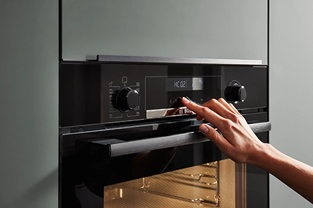 Eine Hand progrmamiert einen Backofen. Der Backofen ist integriert in einen Hochschrank mit grüner, matter Front und durchgehendem Griff. Eine Hand progrmamiert einen Backofen. Der Backofen ist integriert in einen Hochschrank mit grüner, matter Front und durchgehendem Griff.