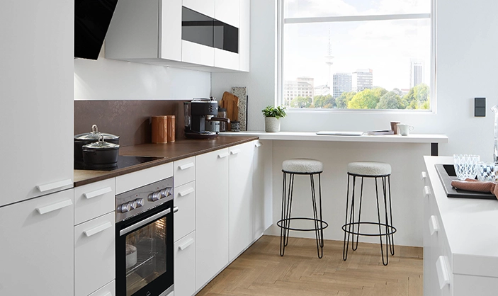 Weiße Küchenzeile mit Buffettlösung an gegenüberliegender Wand und Tresen mit Barhockern am zentralen Fenster. Frontne und Griffe sind mattweiß, die Arbeitsplatte in einem Holzton. Minimalistische Küchenplanung. Weiße Küchenzeile mit Buffettlösung an gegenüberliegender Wand und Tresen mit Barhockern am zentralen Fenster. Frontne und Griffe sind mattweiß, die Arbeitsplatte in einem Holzton. Minimalistische Küchenplanung.