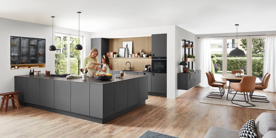 Eine Mutter und ihre Tochter stehen lächelnd in einer modernen, offenen Küche mit einer großen, grifflosen Kochinsel in mattem Dunkelgrau. Die Küche hat Holzakzente und geht in einen Essbereich mit Blick in den Garten über.
