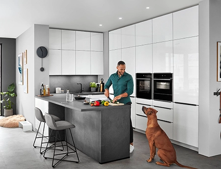 Junger Mann mit Hund in einer modernen weißen Hochglanzküche. Die Küche ist grifflos, sie hat eine Eckform, davor eine Kücheninsel in betongrau. Junger Mann mit Hund in einer modernen weißen Hochglanzküche. Die Küche ist grifflos, sie hat eine Eckform, davor eine Kücheninsel in betongrau.