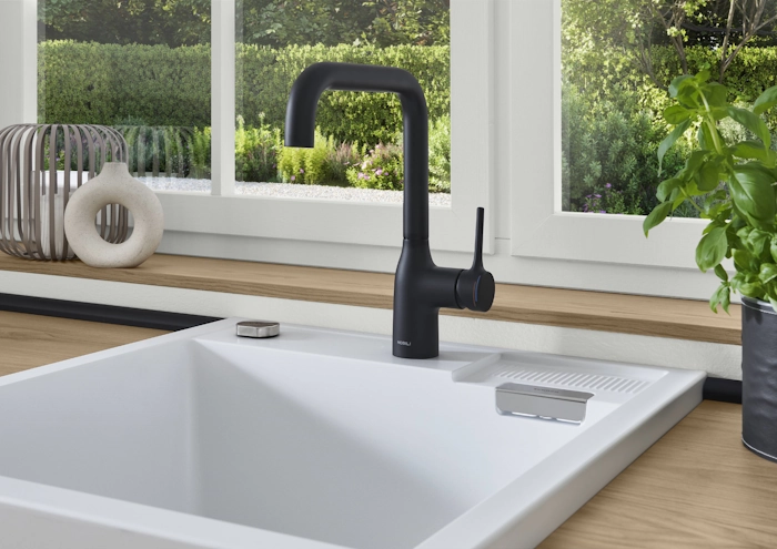 Nahaufnahme einer weißen Spüle in einer Landhausküche mit schwarzer Armatur, Holzarbeitsplatte und Blick ins Grüne durch das Fenster. Nahaufnahme einer weißen Spüle in einer Landhausküche mit schwarzer Armatur, Holzarbeitsplatte und Blick ins Grüne durch das Fenster.