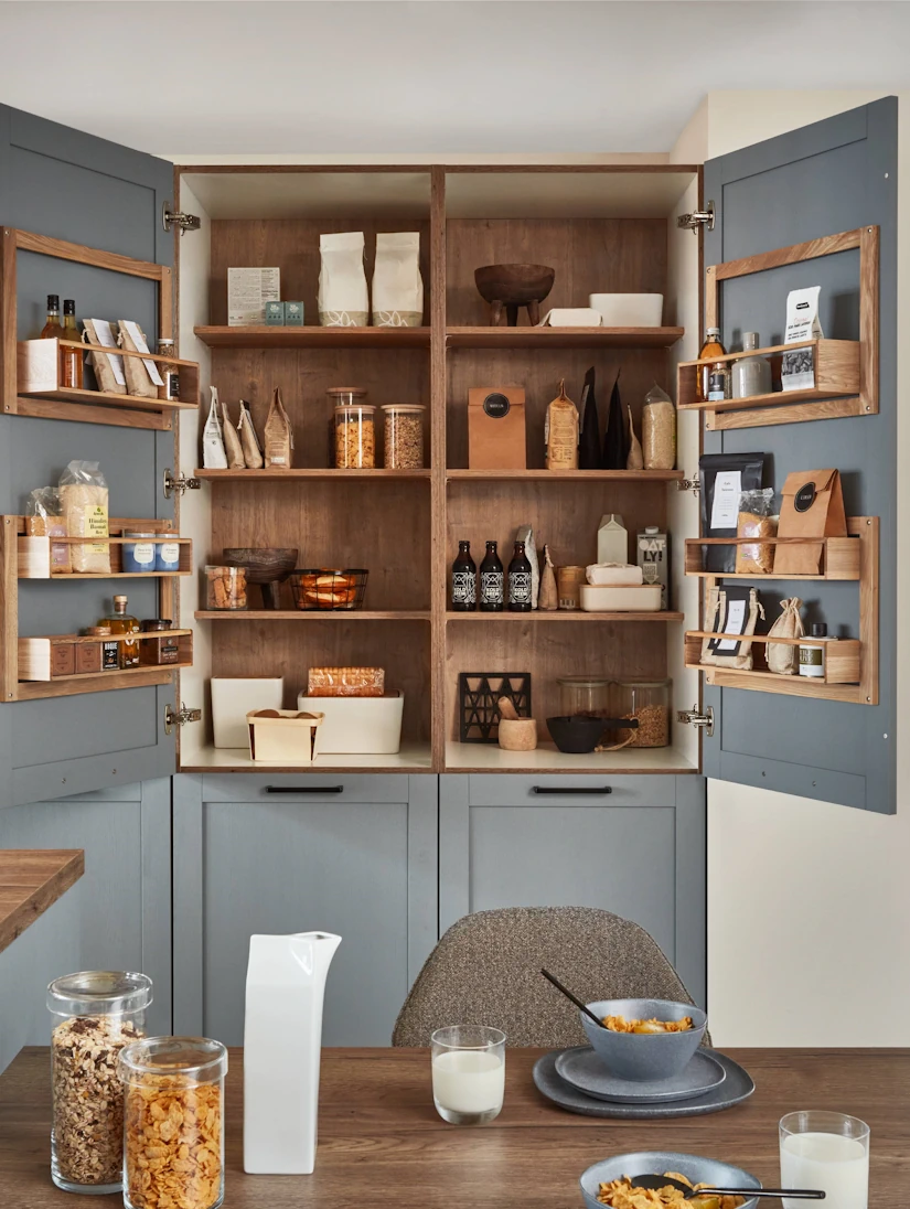 Offener Vorratsschrank in einer Landhausküche mit Holzregalen, gefüllt mit Lebensmitteln. Im Vordergrund steht ein gedeckter Frühstückstisch mit Milch und Müslischalen.