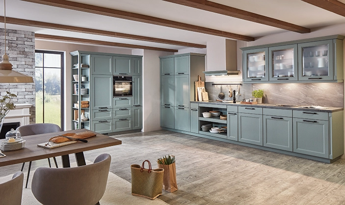 Offene Küche im mediterranen Stil mit salbeigrünen Rahmenfronten. Die Wandschränke sind in Vitrinenausführung, im Vordergrund sehen wir einen Esstisch Offene Küche im mediterranen Stil mit salbeigrünen Rahmenfronten. Die Wandschränke sind in Vitrinenausführung, im Vordergrund sehen wir einen Esstisch