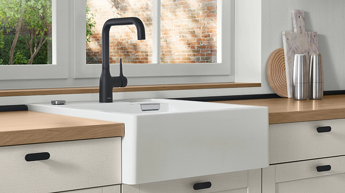 Weiße Landhaus-Küchenspüle mit schwarzer Armatur, Holzarbeitsplatte und Accessoires vor einem Fenster mit Blick auf eine Backsteinwand. Weiße Landhaus-Küchenspüle mit schwarzer Armatur, Holzarbeitsplatte und Accessoires vor einem Fenster mit Blick auf eine Backsteinwand.