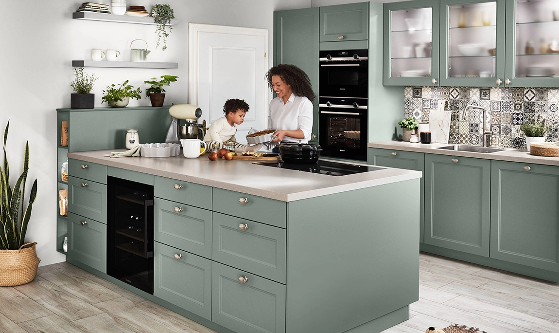 Offene, mediterrane Küche mit grünen Kassettenfronten. Eine junge Mutter zeigt dem Kleinkind eine gefüllte Kuchenform. Offene, mediterrane Küche mit grünen Kassettenfronten. Eine junge Mutter zeigt dem Kleinkind eine gefüllte Kuchenform.