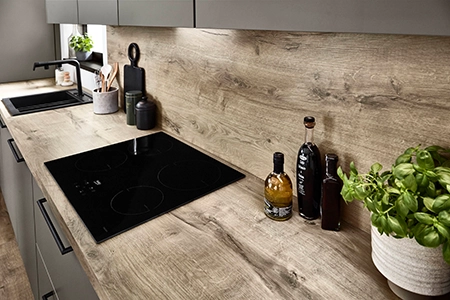 Arbeitsplatte in Holzdekor mit schwarzer Herdplatte und Spüle. Nischenwandverkleidung in gleichem Holzton. Wir sehen Essigflaschen sowie Kräuterpflanzen. Arbeitsplatte in Holzdekor mit schwarzer Herdplatte und Spüle. Nischenwandverkleidung in gleichem Holzton. Wir sehen Essigflaschen sowie Kräuterpflanzen.