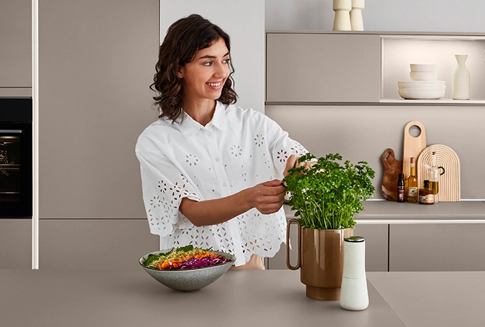 Eine junge Frau in einer Küche im modern natural Stil pflückt Blätter von einer Kräuterpflanze. Sie hat eine weiße Bluse an, die Küche ist grifflos. Eine junge Frau in einer Küche im modern natural Stil pflückt Blätter von einer Kräuterpflanze. Sie hat eine weiße Bluse an, die Küche ist grifflos.